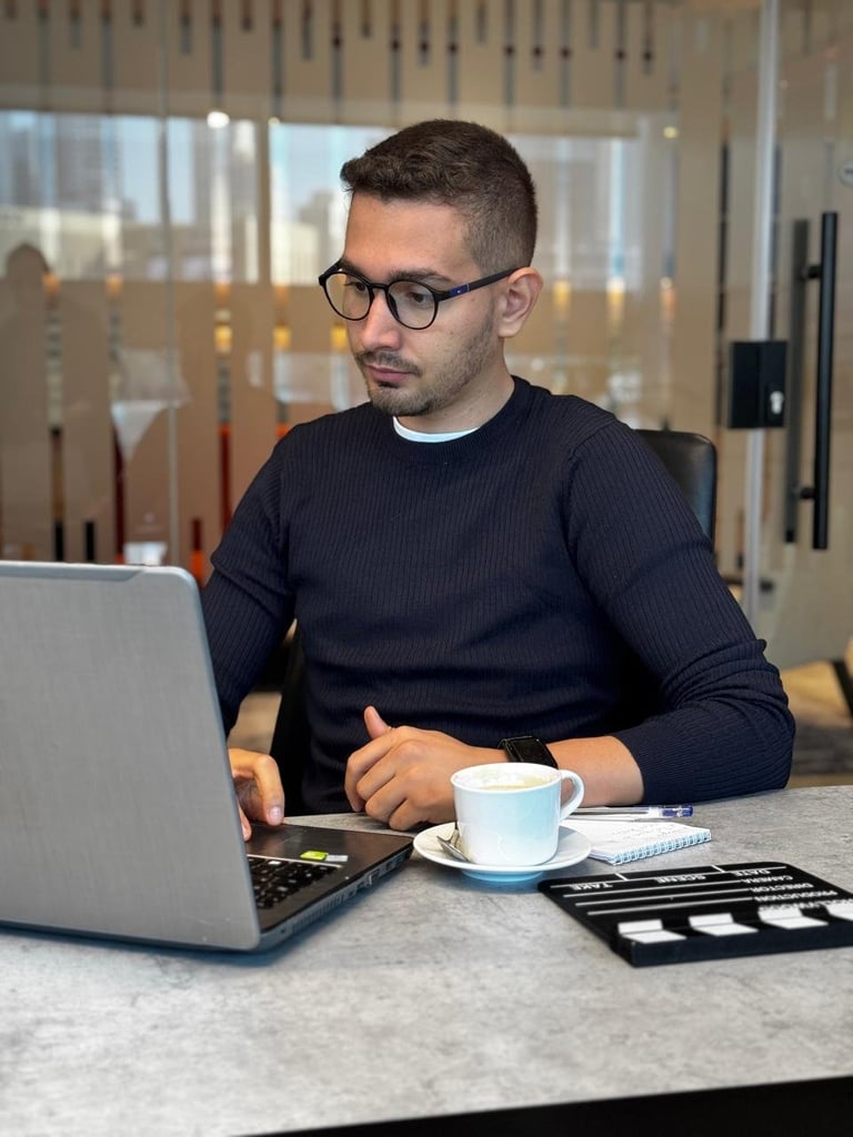 Man wearing glasses and dark sweater working at laptop with coffee cup at desk in modern office
