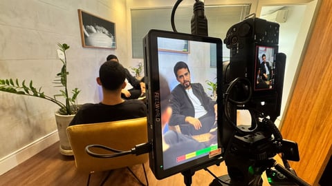 Person in yellow chair being recorded on large monitor displayed on tripod in studio setting with lighting equipment